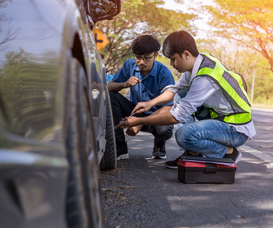 Two men kneel beside a car on a sunny road. One, in a safety vest, uses tools from a red toolbox, while the other watches closely.