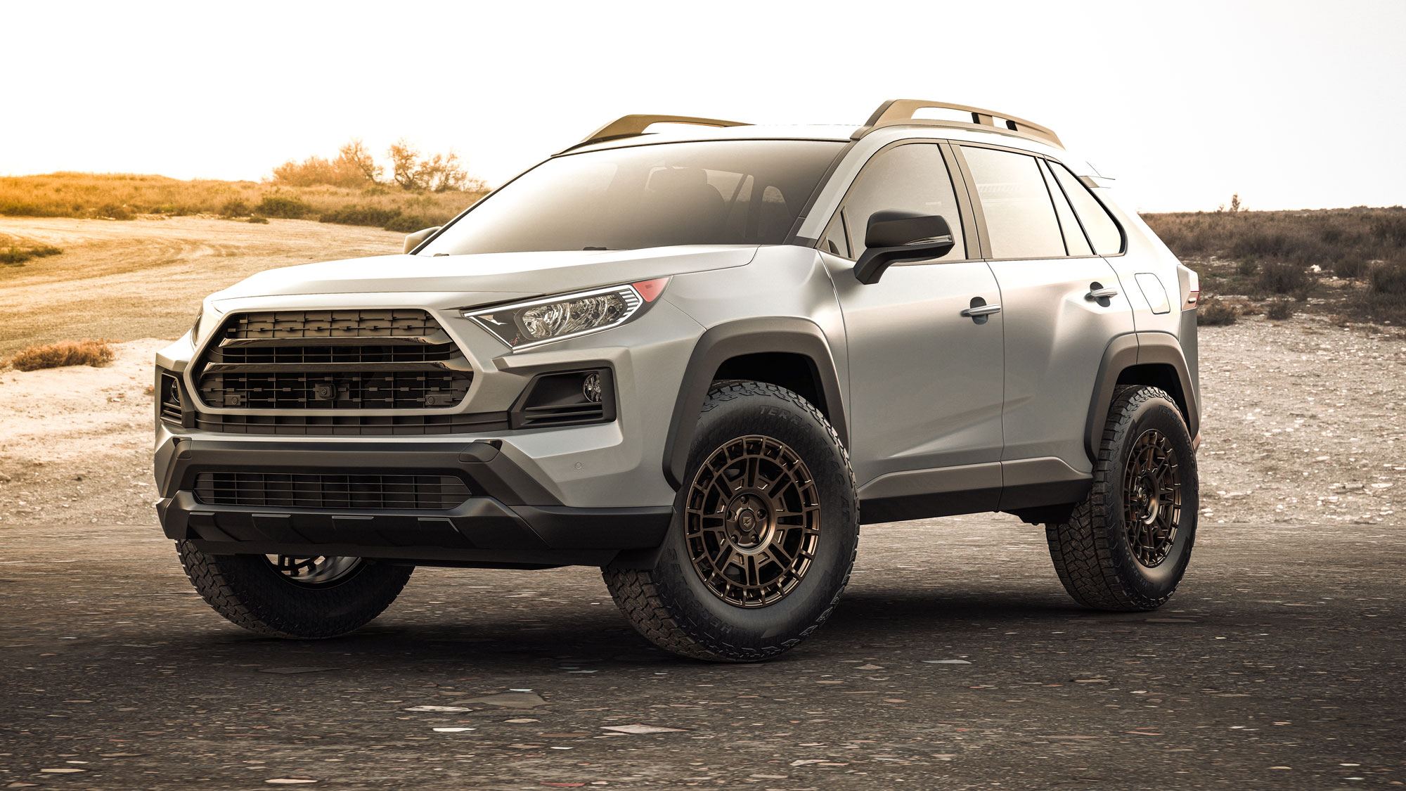 A light gray SUV with black side mirrors and roof rails skewed at an angle from the driver's side. It is parked on a dirt road in an open, barren landscape with sparse vegetation in the background and is equipped with Hercules Terra Trac® AT X-Journey tires.