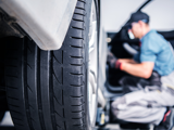 A close-up view of a car tire, focusing on the tread pattern, with a mechanic in the background working on the car's interior. The mechanic is slightly blurred, wearing a cap, gloves, and a blue shirt.