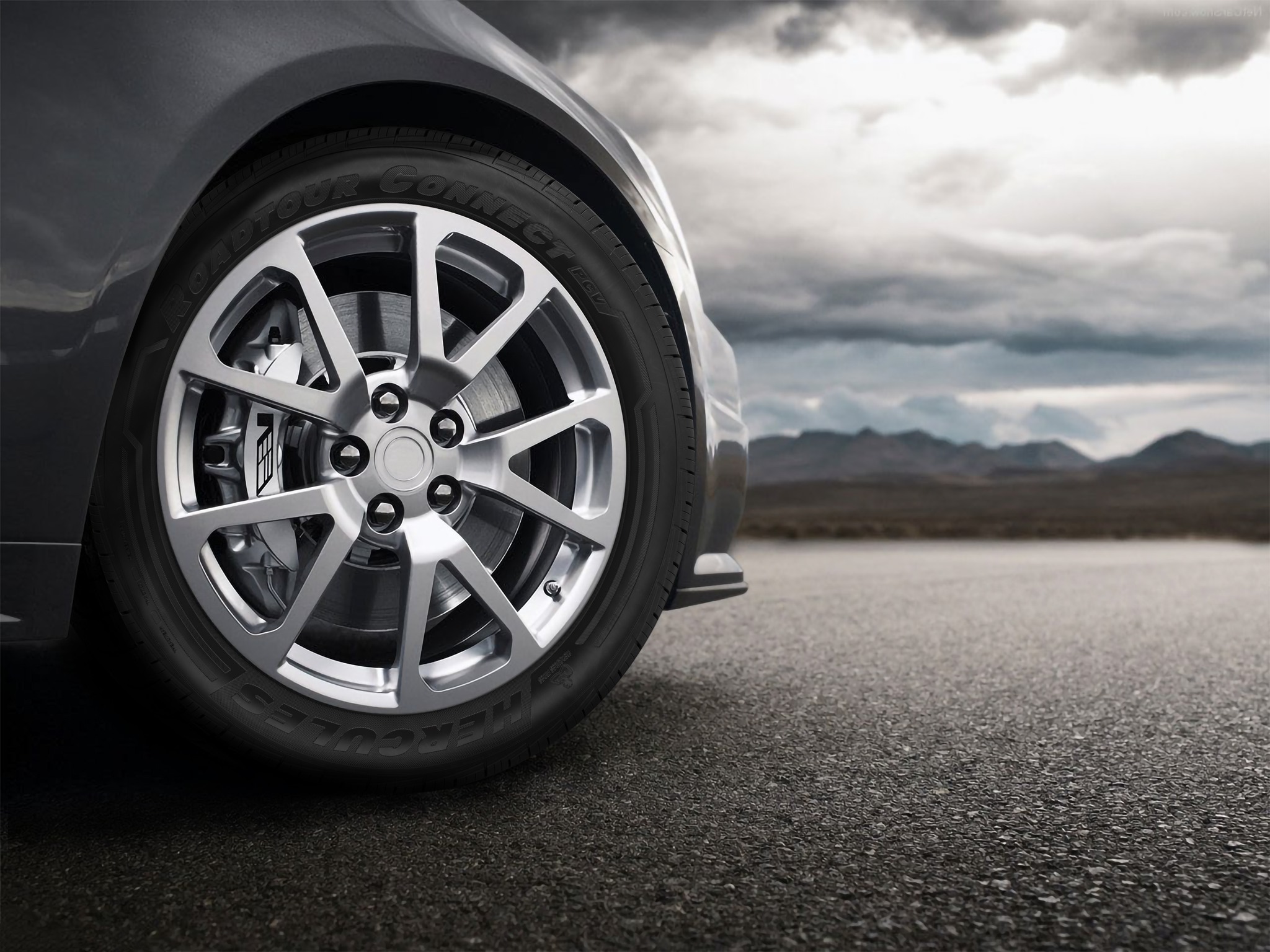 A close-up of a Hercules Tires Roadtour Connect® PCV tire on the passenger side of a dark gray vehicle with a background of mountains on a gloomy day.