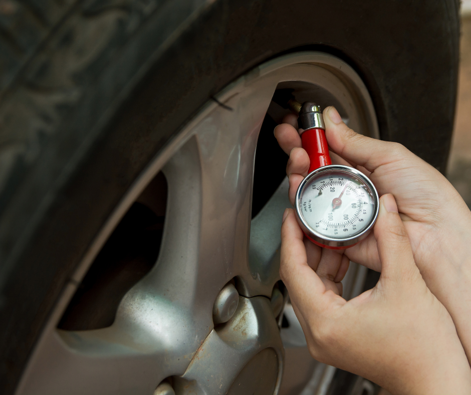 Close-up of hands using a red tire pressure gauge with a round dial display, checking the air pressure in a car tire.