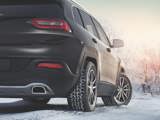 A sleek black SUV parked on a snowy road, showcasing winter tires with a tread pattern designed for traction on icy surfaces. The background features snow-covered trees under a soft, warm sunrise or sunset glow.