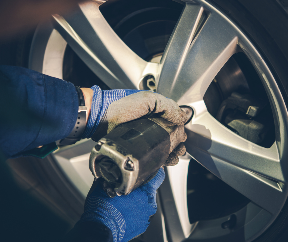 A close-up of two hands wearing blue maintenance gloves, removing the lug nuts off of a tire.