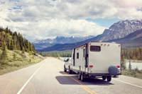 A white pickup truck is towing a large white travel trailer along a scenic highway. The road is flanked by green pine trees on the left and a river on the right. In the background, there are  snow-capped mountains under a partly cloudy sky. 