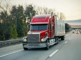  An 18-wheeler semi truck with a red cab on a highway driving in the right lane with passenger vehicles behind it.