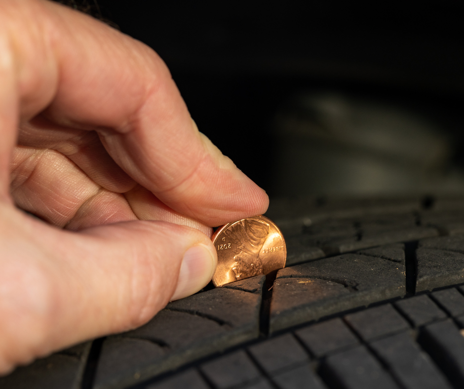 A hand is holding a penny face up with President Lincoln's head upside down being inserted into a tire's tread.